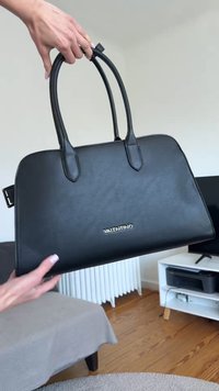 Μαύρη δερμάτινη τσάντα Valentino που κρατιέται με δύο χέρια με στρογγυλές λαβές σε ένα μοντέρνο σαλόνι.