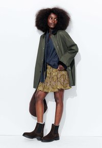 Veste vert olive, chemise en denim bleu, jupe à motifs jaunes, bottines en suède marron, tenant un sac marron, cheveux texturés.