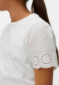 Mujer joven con cabello oscuro en una coleta que lleva una blusa blanca con detalles de encaje bordado en las mangas cortas abullonadas.