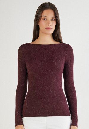Femme aux longs cheveux foncés portant un haut ajusté à manches longues, bordeaux tacheté, et un pantalon blanc, debout devant un fond clair uni.