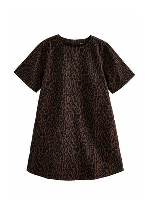 Mujer con un vestido corto oscuro con estampado de leopardo y zapatos negros, de pie junto a una pared de ladrillos rojos, llevando un bolso negro de bandolera.