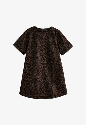 Mujer con un vestido corto oscuro con estampado de leopardo y zapatos negros, de pie junto a una pared de ladrillos rojos, llevando un bolso negro de bandolera.