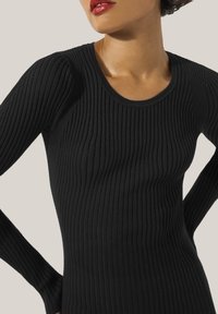 Schwarzes, geripptes Langarmoberteil mit rundem Ausschnitt, das aus einem strukturierten Stoff besteht und eine figurbetonte Silhouette bietet. Sanfter Abschluss auf dem Hautton.