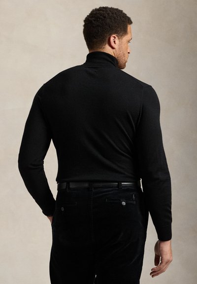 Άνδρας που φοράει μαύρο πουλόβερ με ψηλό γιακά και μαύρο παντελόνι, γυρισμένος με την πλάτη προς απλό μπεζ φόντο.