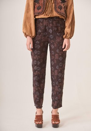 Mujer con pantalones oscuros de cintura alta con estampado y bolsillos parche, combinados con blusa marrón con estampado y sandalias marrones con plataforma.