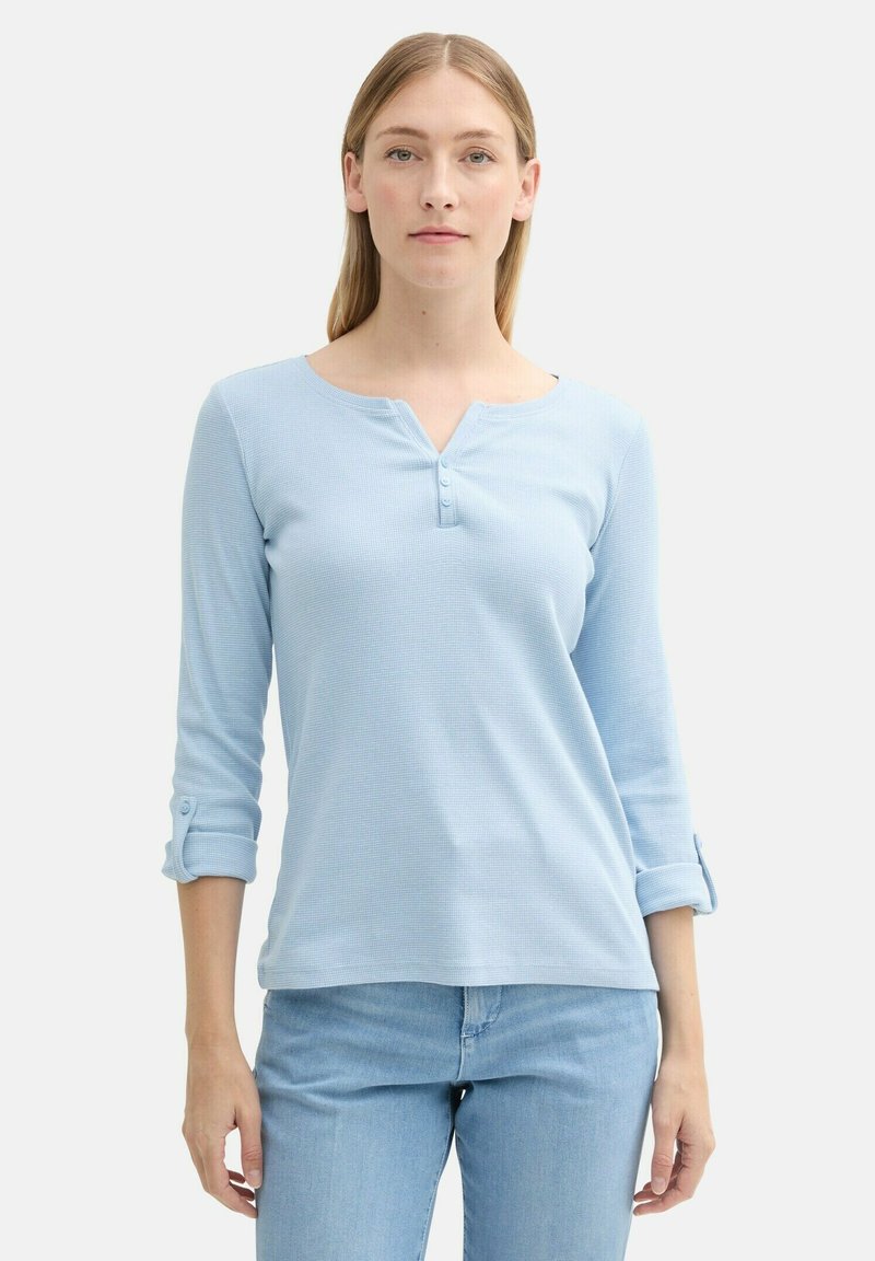 Camisa de manga larga azul claro con escote en V, tres botones en el frente y puños enrollados. Tejido texturizado con un patrón sutil.