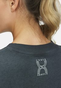 Camiseta de color gris oscuro con cuello redondo y mangas cortas, que presenta un logo y diseño en blanco de "Under Armour" en la parte posterior. Textura suave.