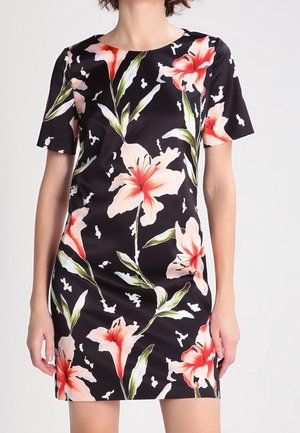 Robe courte noire avec de grandes fleurs de lys roses et rouges et des feuilles vertes, portée par une personne avec les bras détendus sur les côtés.