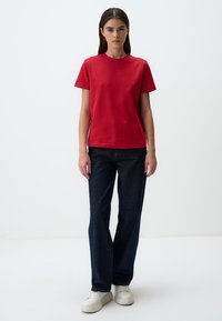 T-shirt rossa di cotone con maniche corte e collo rotondo, abbinata a jeans blu scuro a gamba dritta e sneakers bianche. Design semplice.