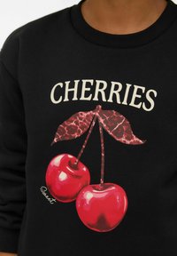 Melns džemperis ar divu sarkanu ķiršu attēlu zaļām lapām un tekstu "CHERRIES" izteiksmīgā krēmkrāsas fontā virs dizaina.