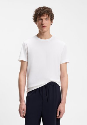 Hombre con cabello rizado que lleva una camiseta blanca lisa de manga corta y pantalones negros con cordón, de pie frente a un fondo blanco.