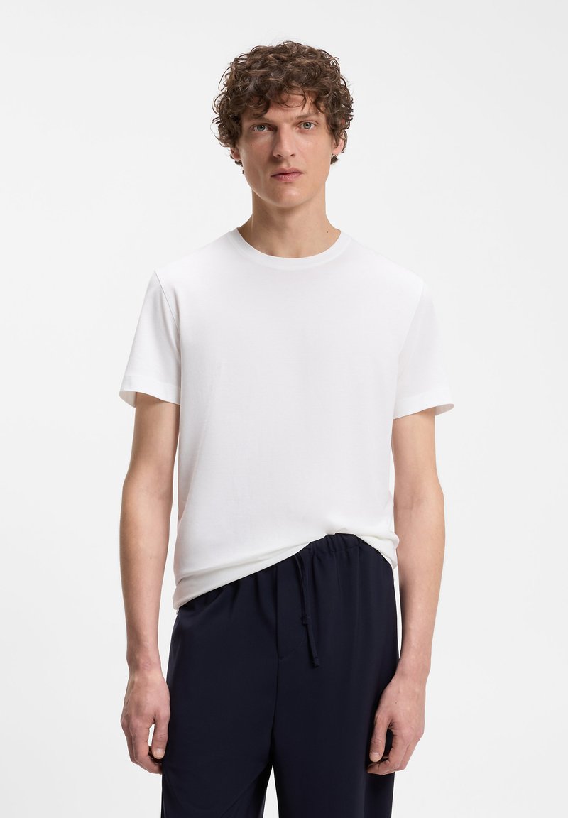 Homme aux cheveux bouclés portant un t-shirt blanc uni à manches courtes et un pantalon noir à cordon, debout devant un fond blanc.