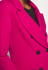 Fuchsiové sako s dvouřadým zapínáním, černými knoflíky, klopami s výstřihem a hladkou texturou. Na levé straně je viditelná kapsa.