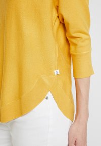 Gelber Strickpullover mit Dreiviertelärmeln, seitlichen Schlitzen und einem leicht verlängerten Rückensaum, getragen über weißen Hosen. Etikett seitlich sichtbar.