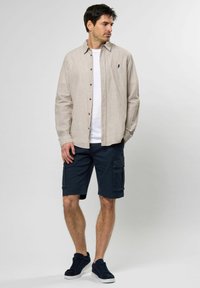 Hellbraune Langarm-Hemdbluse über einem weißen T-Shirt; marineblaue Cargo-Shorts mit seitlichen Taschen; dunkelblaue Sneakers.