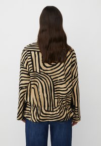 Camicia beige a maniche lunghe con linee ondulate nere, vestibilità ampia con dettaglio vita arricciato e orlo arrotondato. È visibile un abbinamento con denim.