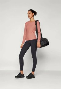 Woman wearing a long-sleeve pink top, black leggings, and black sneakers, carrying a black gym bag over her shoulder, standing against a white background.
