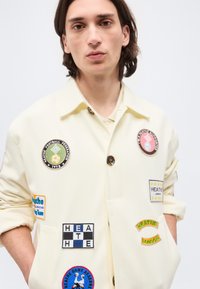 Νεαρό άτομο με καστανά μαλλιά μέχρι τους ώμους φοράει εκρού μπουφάν διακοσμημένο με πολλά πολύχρωμα μπαλώματα και με τα μανίκια γυρισμένα, με τα χέρια στις τσέπες.