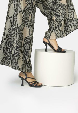 Pies con sandalias negras de tacón alto y tiras, un pie sobre un pedestal cilíndrico blanco, vistiendo pantalones transparentes con patrones florales negros.