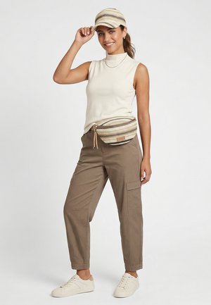 Woman smiling, wearing striped cap, cream sleeveless top, khaki cargo pants, matching striped fanny pack, and white sneakers, standing on white background.