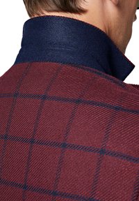 Wełniana marynarka w ciemnym burgundzie z granatowym filcowym kołnierzykiem. Posiada fakturowaną tkaninę z delikatnym wzorem w kratę i kontrastującymi szwami.