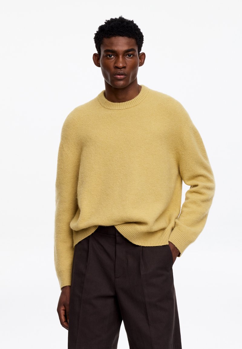 Homme portant un pull ample marron clair et un pantalon marron foncé, debout avec une main dans la poche devant un fond blanc uni.
