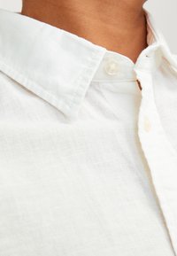 Camicia bianca con colletto realizzata in lino leggero. Presenta una chiusura con bottone e una superficie leggermente testurizzata con design traspirante.