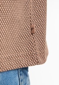 Brown textured fabric featuring a mesh pattern, with a small logo tag attached at the hem. Paired with blue denim at the bottom.