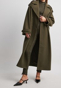 Manteau long vert olive avec un design à double boutonnage, grands revers et manches retroussées. Porté par-dessus une tenue ajustée scintillante et des mules noires à talons.