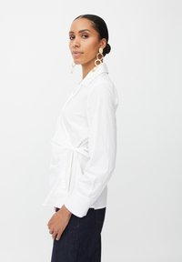 Camisa blanca de manga larga con un diseño envolvente, cintura anudada y cuello clásico. Presenta una tela texturizada y mangas ajustadas, combinada con grandes pendientes de oro.