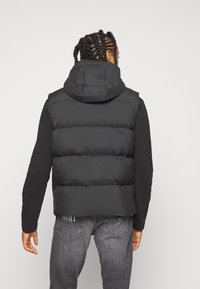 Gilet nero imbottito con cappuccio, caratterizzato da un tessuto texturizzato e cuciture in vista. Indossato sopra una maglietta nera a maniche lunghe e jeans grigi.
