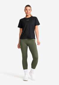 Kvinna som bär en svart kortärmad tröja, olivgröna leggins med logotyp, vita strumpor och vita sneakers, stående mot en vit bakgrund.