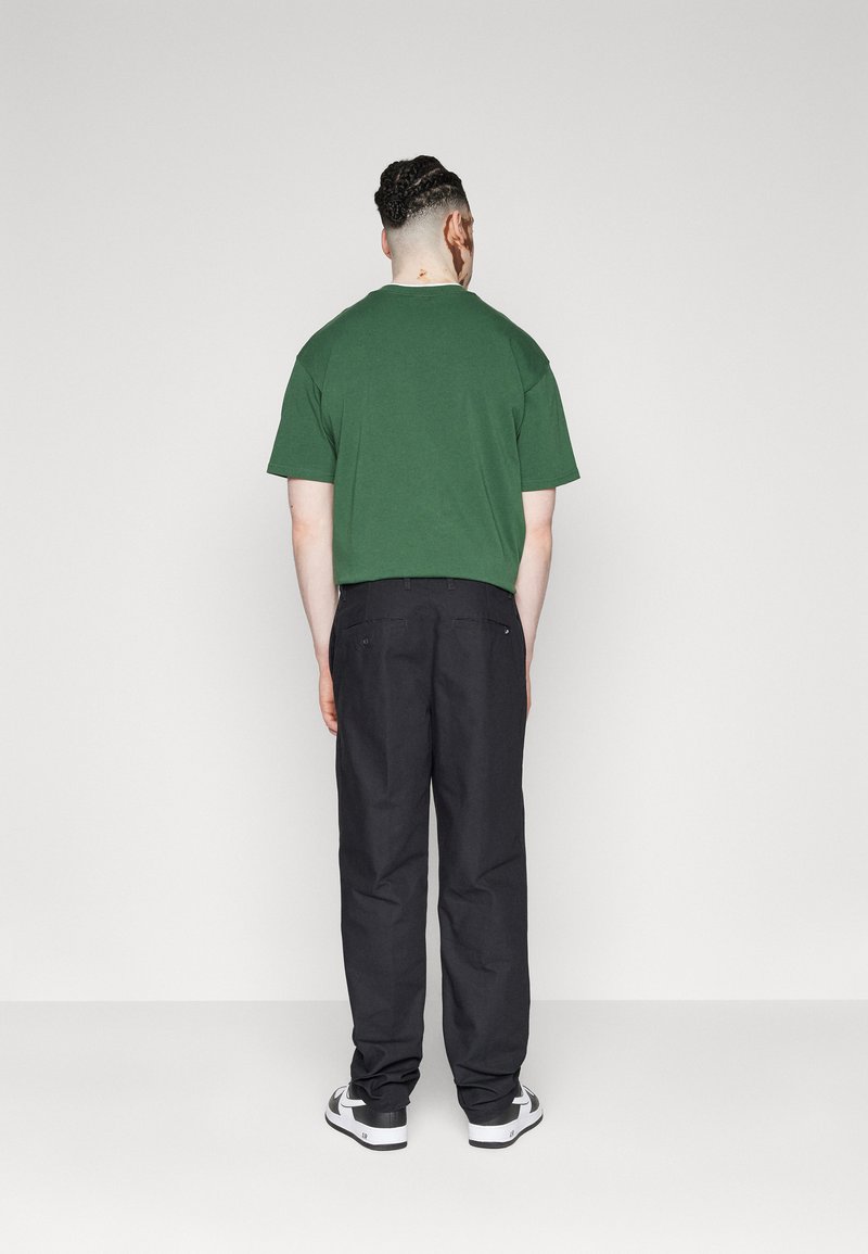 T-shirt vert à manches courtes associé à un pantalon noir, texture du tissu visible. Les chaussures sont blanches avec des accents noirs. Vue de derrière.