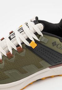 Buty sportowe z teksturowanym oliwkowym cholewką, beżowymi sznurowadłami, czarnymi akcentami i wytrzymałą podeszwą z kontrastującymi pomarańczowymi detalami.