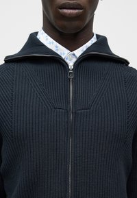 Κοντινό πλάνο ενός ατόμου που φοράει σκούρο μπλε πλεκτό πουλόβερ με φερμουάρ πάνω από ένα λευκό πουκάμισο με μπλε λουλουδάτο μοτίβο.