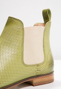 Botín de cuero perforado verde con paneles laterales elásticos beiges y un pequeño tacón. Presenta detalles de costura y una textura suave.