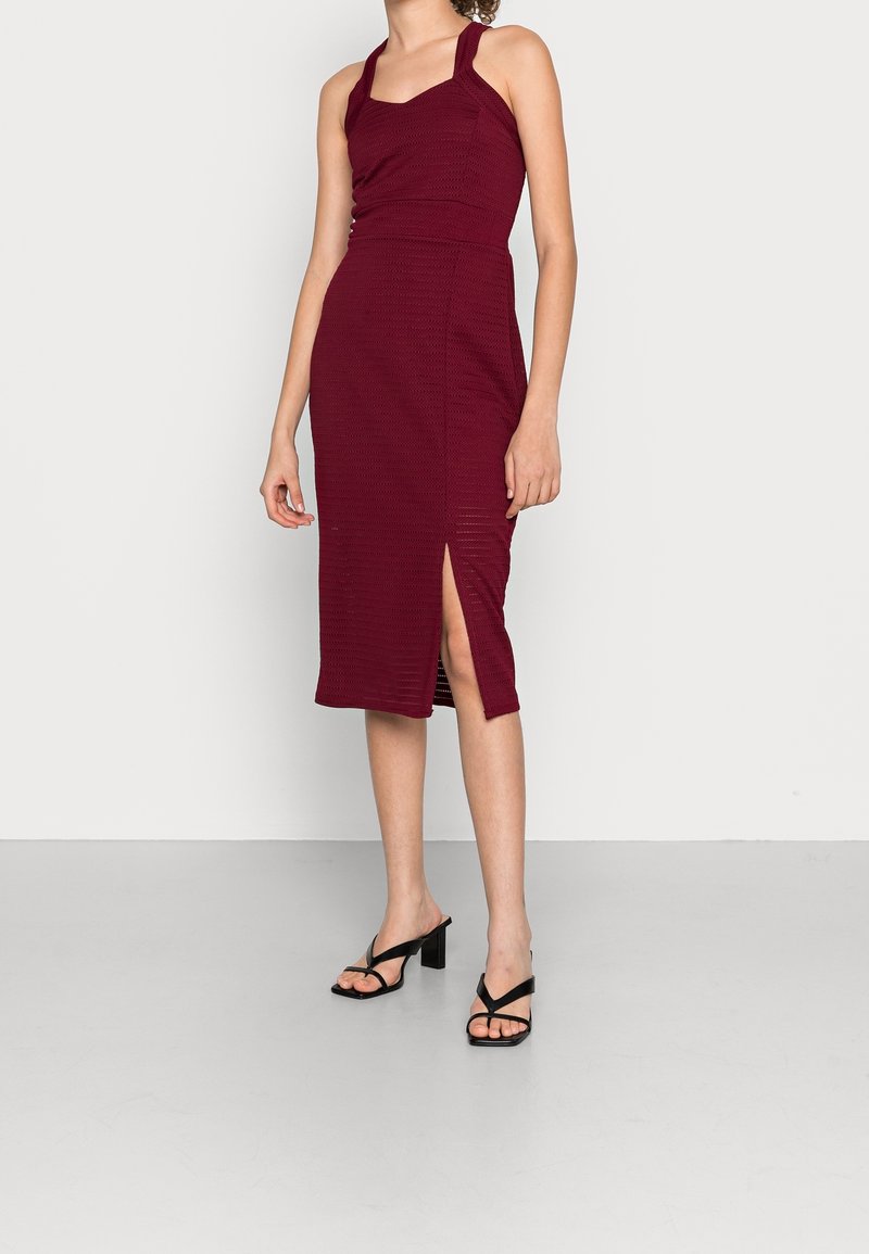 Robe midi bordeaux côtelée avec un décolleté carré et des bretelles halter, dotée d'une fente latérale et d'une silhouette ajustée. Talons ouverts noirs.
