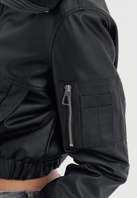 Black leather cropped bomber jacket with a ribbed collar, zippered sleeve pocket, and gathered hem. Smooth texture with a fitted silhouette.