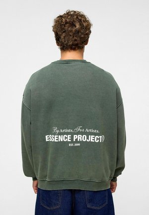 Groene katoenen sweatshirt met een losse pasvorm, geribbelde ronde hals en witte tekst op de rug: "Door kunstenaars, Voor kunstenaars. ESSENCE PROJECT OPGERICHT IN 2000."