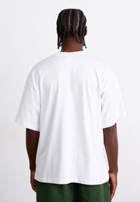 White cotton t-shirt with short sleeves, relaxed fit, and a straight back hem. Viewed from the rear, showcasing minimal design and smooth texture.
