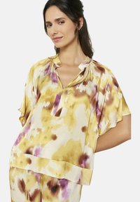 Koszula drukowana w kolorze żółtym, z purpurowymi i brązowymi akcentami. Krótkie rękawy, kołnierzyk mandarynka i miękka, zwiewna tkanina.