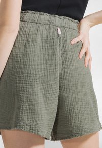 Frau trägt strukturierte olivgrüne Shorts mit elastischem Bund und kleinem pinkfarbenem Etikett, Hand auf der Hüfte ruhend, oberhalb des Bunds ist ein schwarzes Oberteil sichtbar.