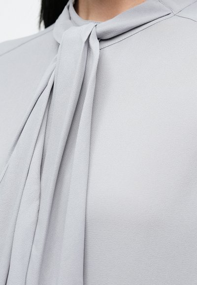 Jasnoszara bluzka zawiązywana przy szyi, pokazująca fakturę tkaniny oraz część szyi i ramienia osoby.