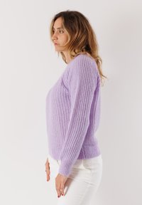 Donna con capelli biondi ondulati, che indossa un maglione di lana lavanda e pantaloni bianchi, in piedi di profilo su uno sfondo bianco uniforme.