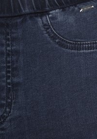 Dark blue denim fabric with a smooth texture, featuring a front pocket and subtle contrasting stitching. Small metallic logo accent present.