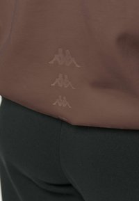 Trois petits logos réfléchissants Kappa empilés verticalement au dos d'un vêtement marron au-dessus d'un pantalon noir.