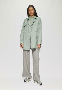 Trench de color verde claro con botones de doble botonadura, llevado sobre una blusa gris texturizada y pantalones anchos de patrón, combinado con zapatillas plateadas.