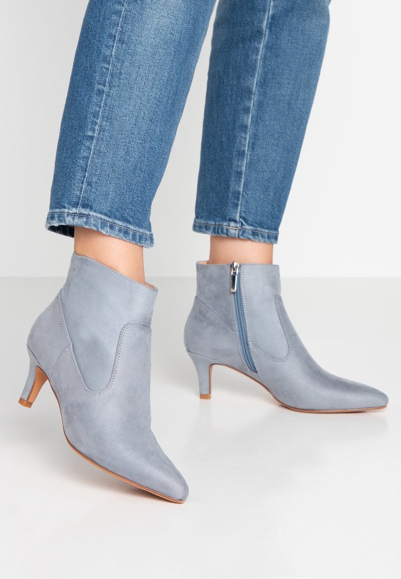 Personne portant des bottines en daim bleu clair à talons kitten et fermetures éclair latérales, associées à un jean bleu raccourci, sur un sol blanc.
