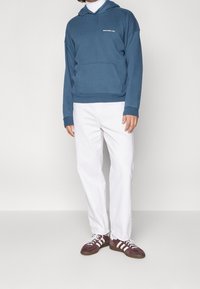 Mann trägt einen blauen Hoodie, einen weißen Rollkragenpullover, weiße Hosen und braune Sneakers mit weißen Streifen und steht vor einem schlichten Hintergrund.