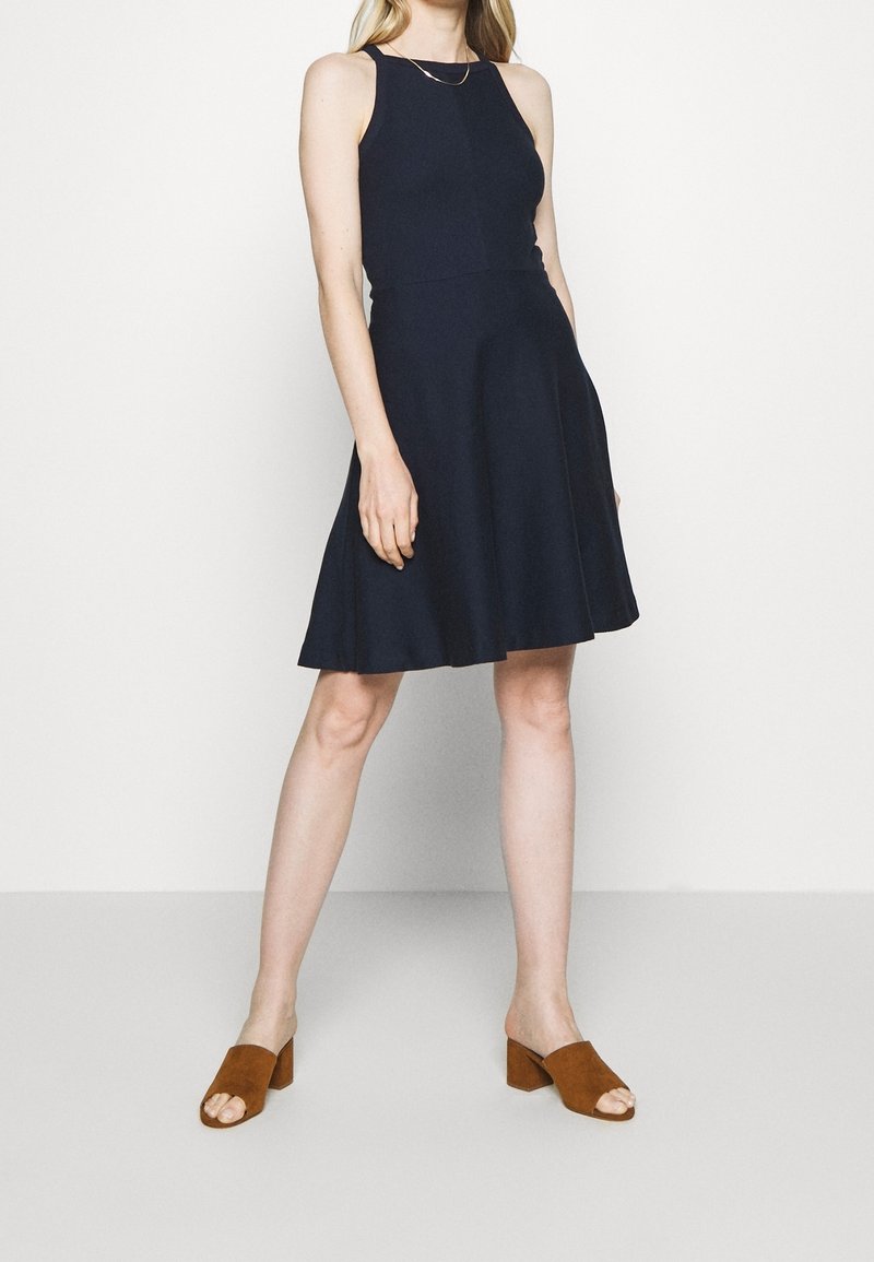 Femme portant une robe noire sans manches longueur genoux et des sandales marron à talons blocs et bouts ouverts, debout devant un fond blanc.